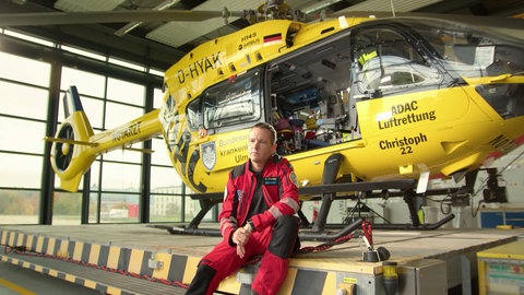Dr. Christian Fischer sitzt in einer roten Uniform vor dem gelben ADAC-Rettungshelikopter Christoph 22 in einem Hangar.