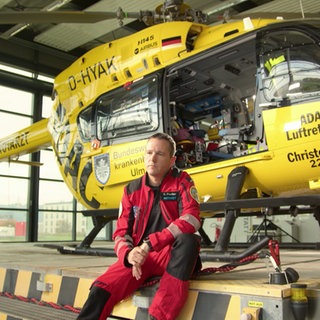 Dr. Christian Fischer sitzt in einer roten Uniform vor dem gelben ADAC-Rettungshelikopter Christoph 22 in einem Hangar.