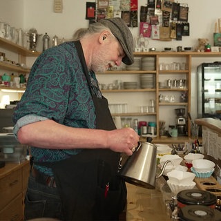 Ein Barista steht an einer hölzernen Theke und schüttet Kaffee aus einer metallenen Kanne in einen Kaffeefilter.