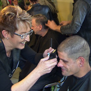 Eine Friseurin der Barber Angels lächelt und schneidet einem sitzenden Mann mit einem Rasierer die Haare.