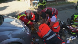Eine Gruppe von in orangen Anzügen gekleideten Sanitätern kniet um eine liegende Person. Rechts von ihnen steht ein Auto, drum herum liegt viel medizinisches Equipment.