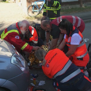 Eine Gruppe von in orangen Anzügen gekleideten Sanitätern kniet um eine liegende Person. Rechts von ihnen steht ein Auto, drum herum liegt viel medizinisches Equipment.