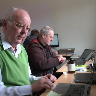 Helmut Petersen sitzt vor einem Laptop und tupft diesen ab. Er trägt dabei einen grünen Pullunder und ein kariertes Hemd. Im Hintergrund sitzen weitere Männer an Laptops und PC-Bildschirmen.