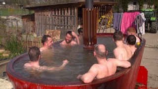 Eine Gruppe von Feuerwehrmännern sitzt oberkörperfrei in einem roten, ehemaligen Futtermischwagen, der mit Wasser gefüllt ist.