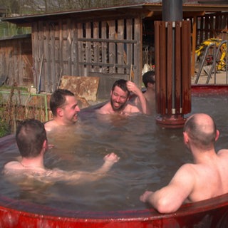 Eine Gruppe von Feuerwehrmännern sitzt oberkörperfrei in einem roten, ehemaligen Futtermischwagen, der mit Wasser gefüllt ist.
