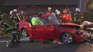 Eine Gruppe von Feuerwehrleuten, Sanitätern und Notärzten steht um ein kaputtes, rotes Auto. Es ist Dämmerung