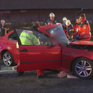 Eine Gruppe von Feuerwehrleuten, Sanitätern und Notärzten steht um ein kaputtes, rotes Auto. Es ist Dämmerung