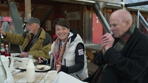 Sonja Faber-Schrecklein sitzt an einem Tisch. Links und rechts von ihr sitzen zwei Männer. Auf dem Tisch stehen Kuchen und Getränke