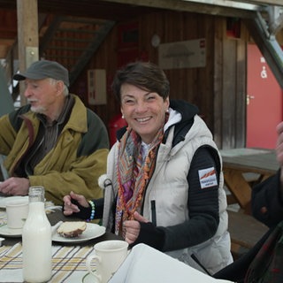 Sonja Faber-Schrecklein sitzt an einem Tisch. Links und rechts von ihr sitzen zwei Männer. Auf dem Tisch stehen Kuchen und Getränke