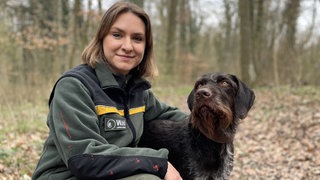 Lia Ternes kniet auf dem Boden im Wald und hat einen Arm um ihren Hund Pax gelegt. Sie trägt eine grüne Jacke.