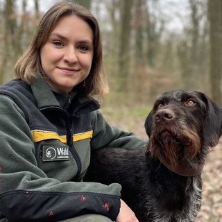 Lia Ternes kniet auf dem Boden im Wald und hat einen Arm um ihren Hund Pax gelegt. Sie trägt eine grüne Jacke.