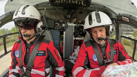 Pilot Jens Jasper und Notfallsanitäter Dirk Hornig sitzen im Cockpit eines Hubschraubers und tragen Pilotenhelme. Jens Jasper hat zudem sein Sonnenvisier runtergeklappt.