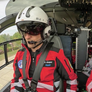 Pilot Jens Jasper und Notfallsanitäter Dirk Hornig sitzen im Cockpit eines Hubschraubers und tragen Pilotenhelme. Jens Jasper hat zudem sein Sonnenvisier runtergeklappt.