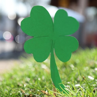 Ein ausgeschnittenes Papier-Kleeblatt steht auf einer grünen Wiese. Im Hintergrund ist unscharf eine Straße zu sehen.