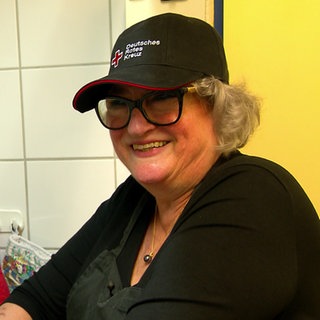 Susanne Gossel steht in vor einer Fliesenwand mit Backofen-Handschuhen. Sie lächelt in die Kamera und trägt eine Mütze mit dem Logo des Deutschen Roten Kreuzes.