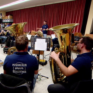 Eine Musikkapelle sitzt in einem Proberaum mit einem roten Vorhang. Im Vordergrund sitzen Menschen mit großen Tubas. Im Hintergrund ist ein Mann zu sehen, der dirigiert.