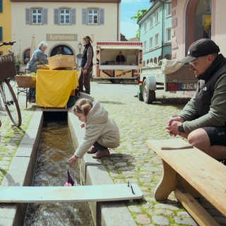 Ein kleines Kind lässt ein Papierboot in einen kleinen Bach in Staufen schwimmen. Links stehen Fahrräder, rechts steht der Vater.