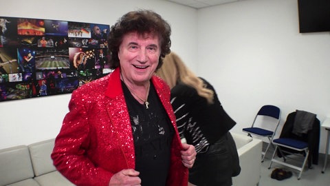 Olaf der Flipper trägt eine rote Anzugsjacke und lächelt in die Kamera. Im Hintergrund steht seine Tochter Pia. Sie stehen in einem weißen Raum in der Mannheimer SAP Arena.