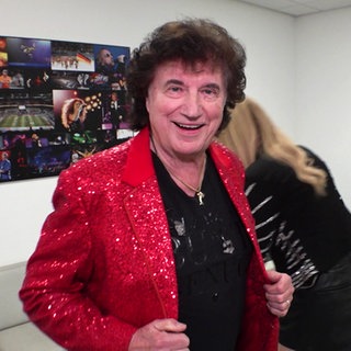 Olaf der Flipper trägt eine rote Anzugsjacke und lächelt in die Kamera. Im Hintergrund steht seine Tochter Pia. Sie stehen in einem weißen Raum in der Mannheimer SAP Arena.