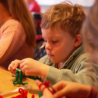 Ein Junge mit blonden Haaren häkelt grüne Wolle mit einer Häkelnadel. Links und rechts von ihm sitzen jeweils zwei Mädchen, die verschwommen sind.