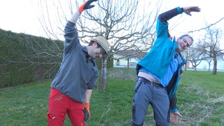 Meinrad und Klaus stehen im Garten und dehnen sich. Sie haben dabei ihre rechte Hand über dem Kopf und neigen sich zur Seite. Beide lachen dabei und tragen Garten-Ausrüstung.