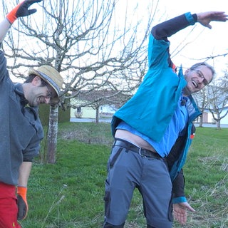 Meinrad und Klaus stehen im Garten und dehnen sich. Sie haben dabei ihre rechte Hand über dem Kopf und neigen sich zur Seite. Beide lachen dabei und tragen Garten-Ausrüstung.