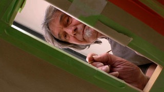 Man sieht Manne Gelmar durch ein viereckiges Loch in einer Holzplatte. Der Mann mit grauen Haaren und Spitzbart hält einen Pinsel in der Hand und malt damit auf der anderen Seite des Brettes.