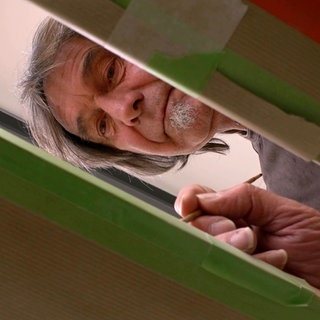 Man sieht Manne Gelmar durch ein viereckiges Loch in einer Holzplatte. Der Mann mit grauen Haaren und Spitzbart hält einen Pinsel in der Hand und malt damit auf der anderen Seite des Brettes.