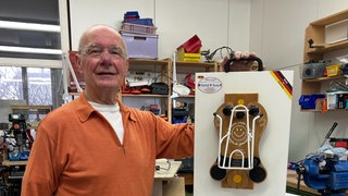 Klaus-Peter Beer steht mit seiner Sockenanziehhilfe in der Hand in einer Werkstatt.