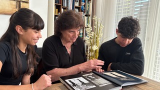 Olaf der Flipper sitzt in einem schwarzen Hemd an einem Tisch und blättert in einem Bilderbuch. Links und rechts von ihm sitzen seine Enkel, die interessiert in das Buch schauen.