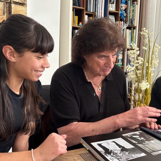 Olaf der Flipper sitzt in einem schwarzen Hemd an einem Tisch und blättert in einem Bilderbuch. Links und rechts von ihm sitzen seine Enkel, die interessiert in das Buch schauen.
