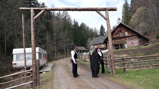 Drei Menschen gehen durch einen hölzernen Torbogen. Links steht ein alter Wohnwagen. Rechts steht ein altes Bauernhaus.
