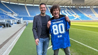Olaf der Flipper steht mit einem blauen Trikot mit der Aufschrift "Karlsruher SC - 80 - Olaf" im Wildparkstadion in Karlsruhe. Neben ihm steht KSC-Geschäftsführer Mario Eggimann.