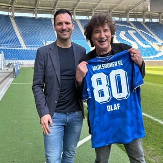 Olaf der Flipper steht mit einem blauen Trikot mit der Aufschrift "Karlsruher SC - 80 - Olaf" im Wildparkstadion in Karlsruhe. Neben ihm steht KSC-Geschäftsführer Mario Eggimann.