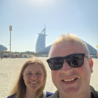 Selfie von Manuela und Ralf Trinkhaus. Sie stehen auf einem Strand, im Hintergrund sieht man das Burj al Arab in Dubai.