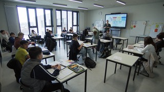 Ein Klassenzimmer in der Oscar-Paret-Schule. Viele Kinder sitzen an weißen Tischen. Im Hintergrund ist ein Mann, der Lehrer, an einem digitalen Whiteboard zu sehen.