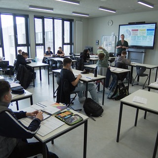 Ein Klassenzimmer in der Oscar-Paret-Schule. Viele Kinder sitzen an weißen Tischen. Im Hintergrund ist ein Mann, der Lehrer, an einem digitalen Whiteboard zu sehen.