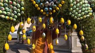 Von einem Baum hängen viele gelbe Ostereier herunter. Hinter diesen Eiern sieht man leicht einen Mann in einer gelben Weste, der die Eier an den Baum hängt.