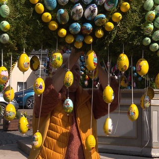 Von einem Baum hängen viele gelbe Ostereier herunter. Hinter diesen Eiern sieht man leicht einen Mann in einer gelben Weste, der die Eier an den Baum hängt.