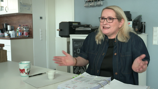 Mechaela, eine 57-jährige, blonde Frau, sitzt an einem Tisch. Vor ihr liegt ein Ordner mit Abrechnungen. Sie hebt ihre Arme und signalisiert Hilflosigkeit.