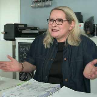 Mechaela, eine 57-jährige, blonde Frau, sitzt an einem Tisch. Vor ihr liegt ein Ordner mit Abrechnungen. Sie hebt ihre Arme und signalisiert Hilflosigkeit.