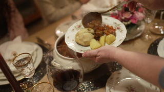 Auf einen Teller wird mit einer Kelle Gulasch geschöpft. Auf dem Teller liegen bereits Kartoffeln und eine Dampfnudel.