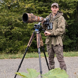 Ein junger Mann in Tarnkleidung steht vor einer Kamera in Tarnfarben. Er ist Tierfotograf.
