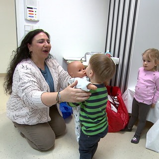 Kursleiterin Songül Demirbile kniet auf dem Boden und übergibt einem kleinen Jungen mit blonden Haaren eine Spielzeug-Puppe.