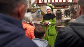 Eine Stadtführerin in grünen Mittelalter-Gewändern steht vor einem Brunnen und redet zu einer Gruppe von Leuten.