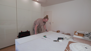 Daniel, ein Mann in Arbeitskleidung und mit blondem Dutt, baut einen Schrank auf.