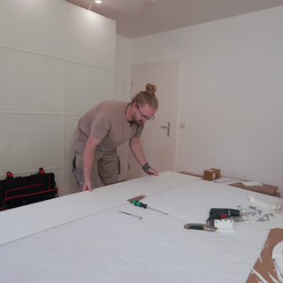 Daniel, ein Mann in Arbeitskleidung und mit blondem Dutt, baut einen Schrank auf.