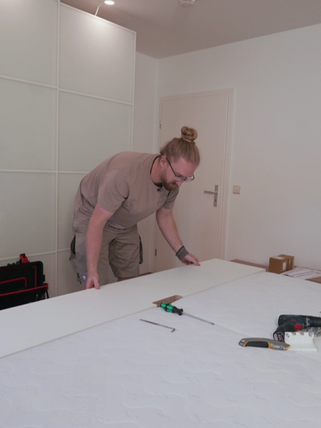 Daniel, ein Mann in Arbeitskleidung und mit blondem Dutt, baut einen Schrank auf.