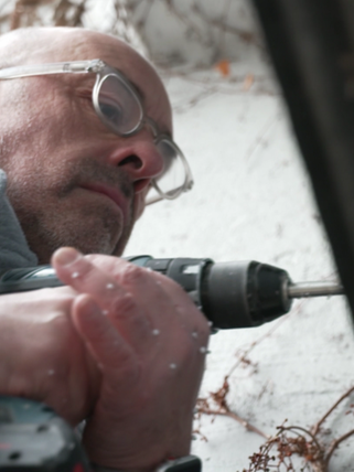 Christian Bauerle steht an einem Garagentor und bohrt mit einem Akkubohrer ein Loch hinein. 