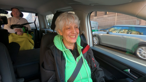 Eine Frau sitzt am Steuer eines Autos. Hinter ihr sitzt ein Mann im Rollstuhl. 